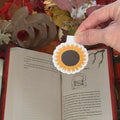 A hand holds a die-cut paper bookmark shaped like a sunflower over an open book on a wooden table, with autumn leaves and fall decorations in the background.