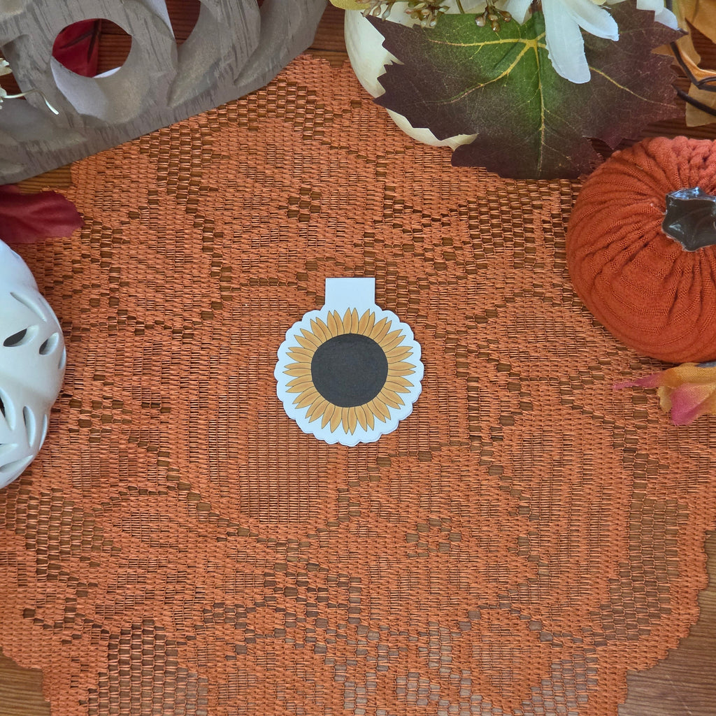 Sunflower magnetic bookmark set on orange doily with autumn decorations in the background.