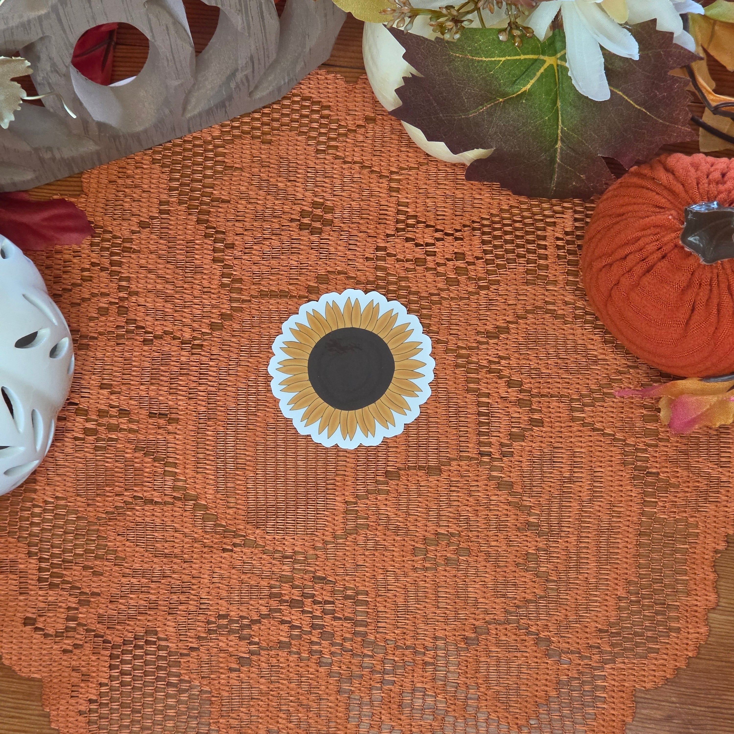 A sticker in the shape of a sunflower sitting on an orange doily with autumn decor in the background.