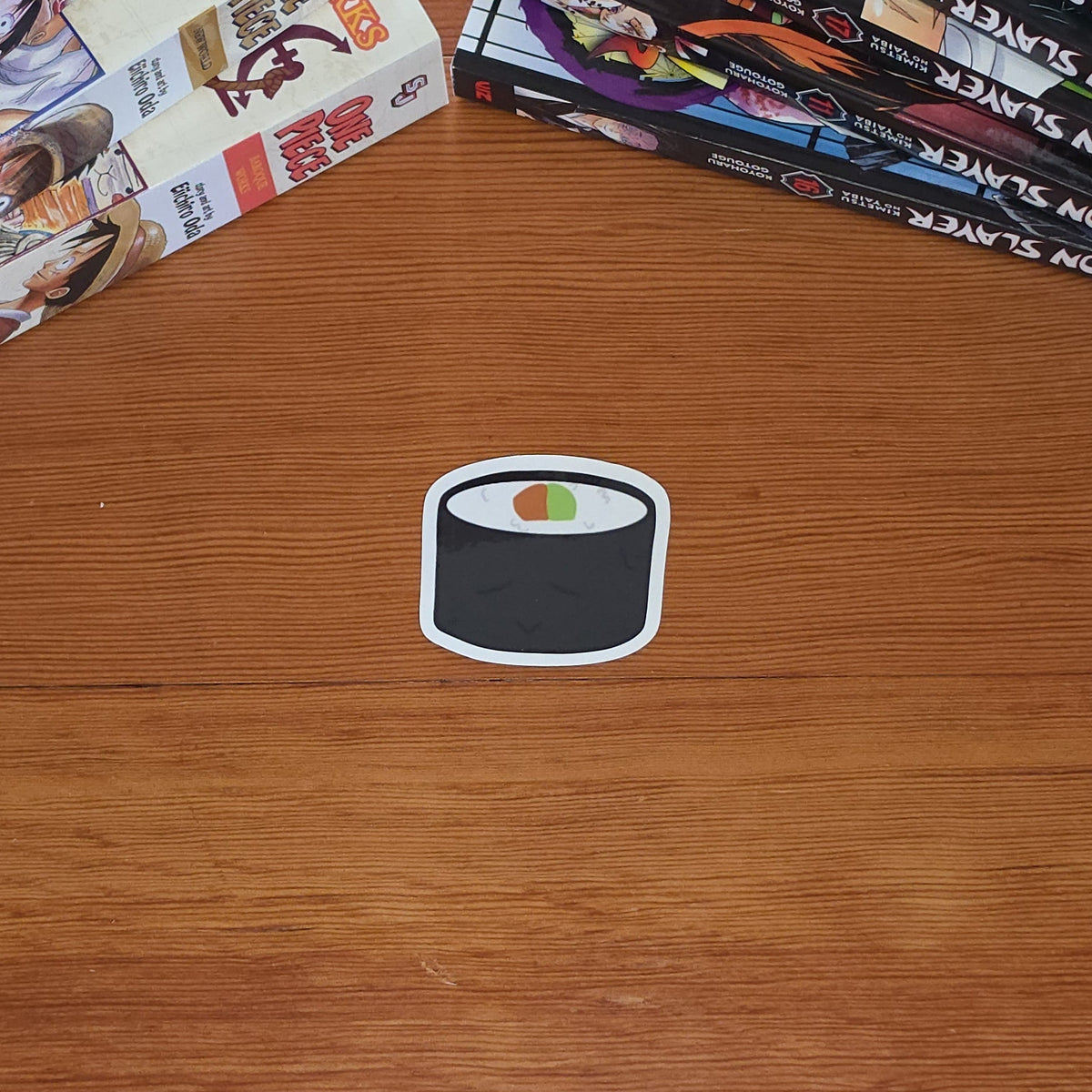 A small die-cut sticker shaped like a sushi roll sits on a wooden tabletop, with stacks of manga books in the background.