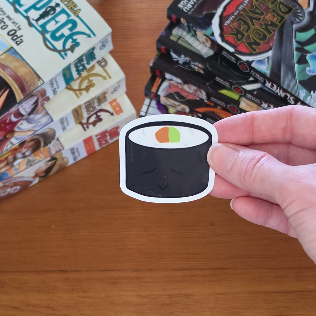 A hand holds a die-cut sticker of a sushi roll with black seaweed, white filling, and a small green and orange accent, over a wooden tabletop with books in the background.