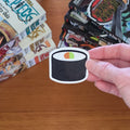 A hand holds a die-cut sticker of a sushi roll with black seaweed, white filling, and a small green and orange accent, over a wooden tabletop with books in the background.