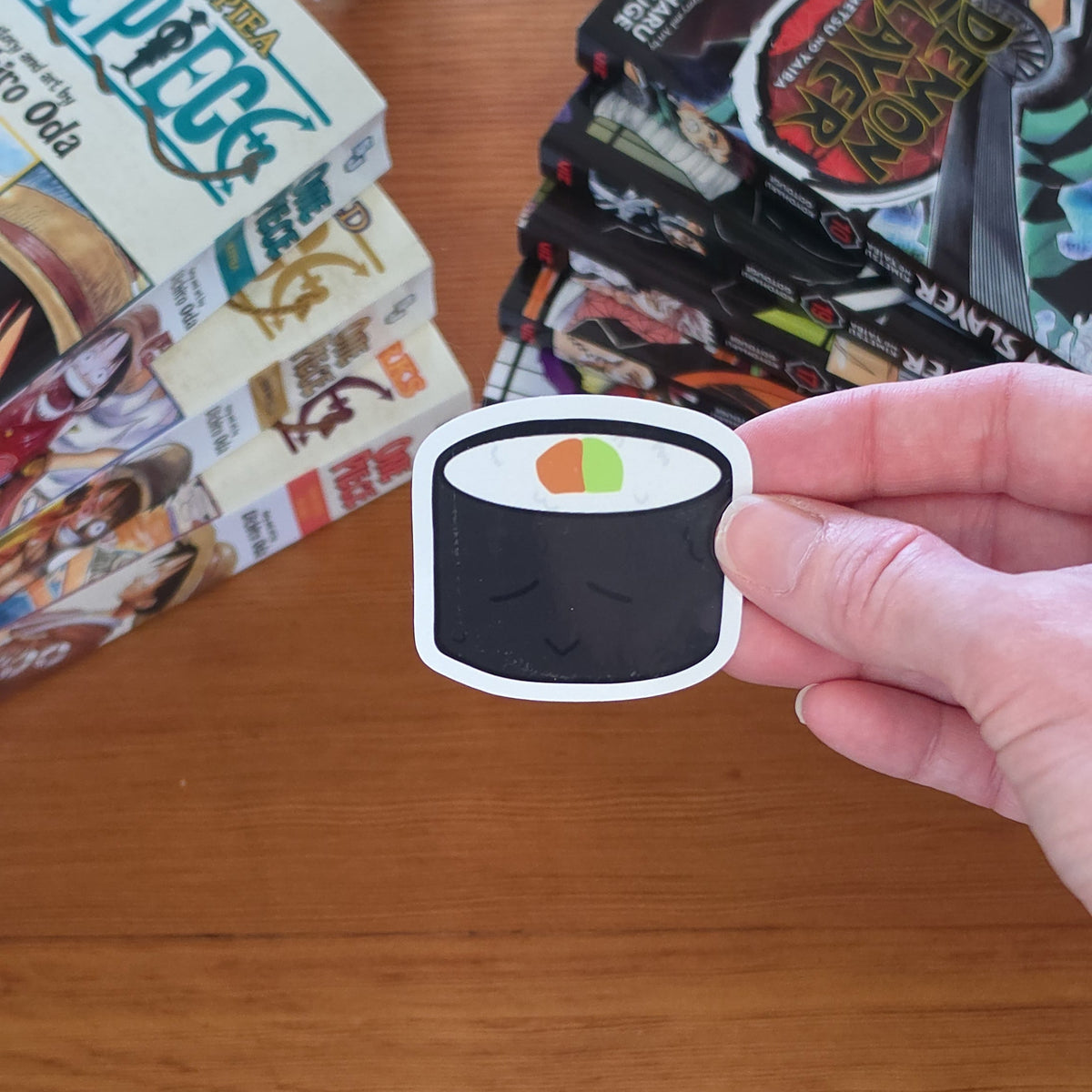 A hand holds a die-cut sticker of a sushi roll with black seaweed, white filling, and a small green and orange accent, over a wooden tabletop with books in the background.