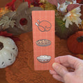 A hand holds an orange patterned bookmark with Thanksgiving-themed illustrations of a turkey, pie, and mashed potatoes, set against a decorated autumn table background.