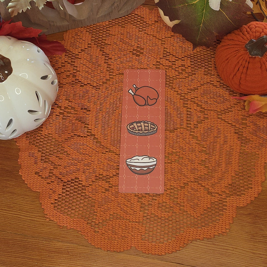 An orange patterned bookmark with Thanksgiving-themed illustrations of a turkey, pie, and mashed potatoes, set against a decorated autumn table background.