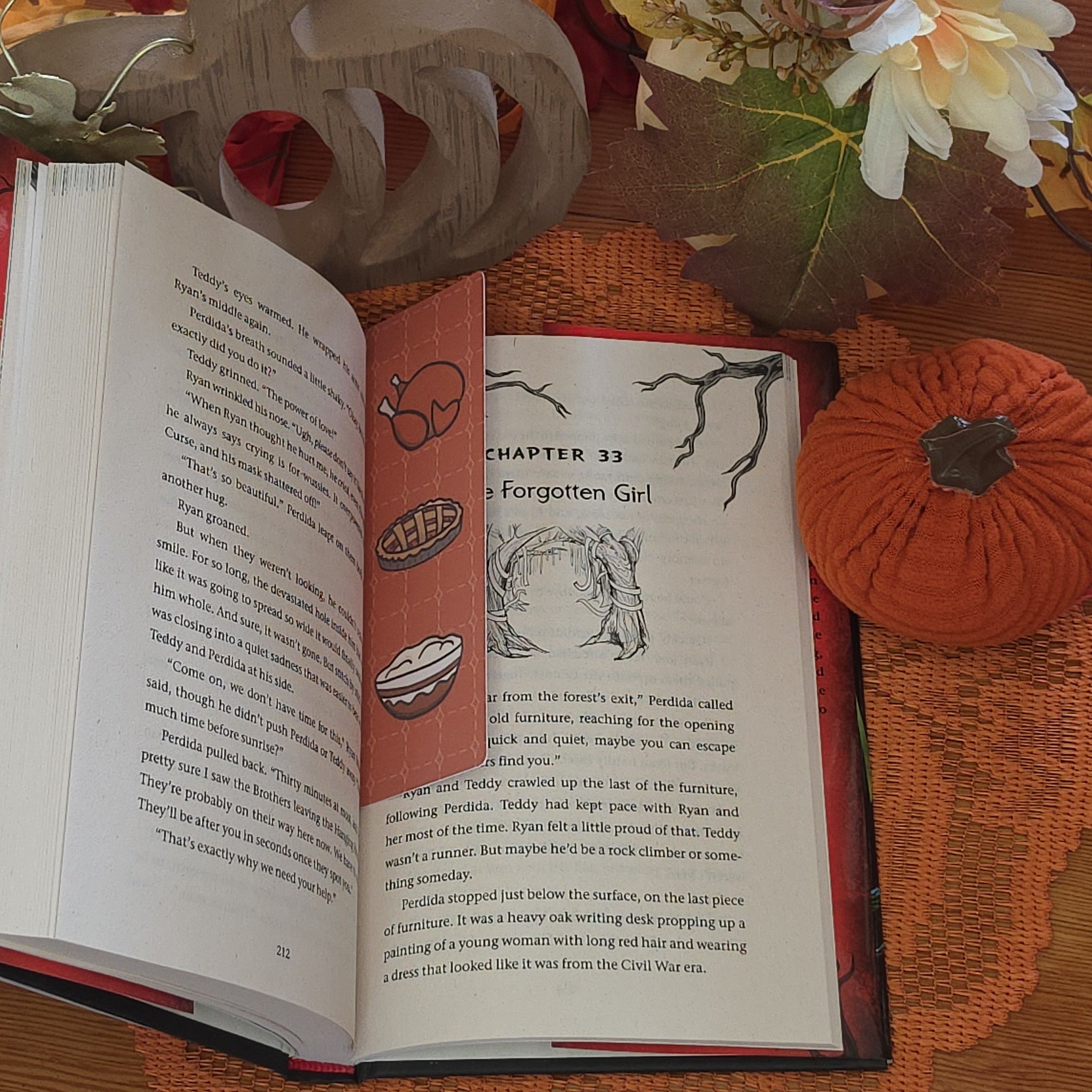 An open book features an orange patterned bookmark with Thanksgiving-themed illustrations of a turkey, pie, and mashed potatoes, set against a decorated autumn table background.