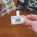 A hand holds a rectangular magnetic bookmark with rounded corners featuring a printed illustration of stacked books and the text 'I own a Thousand books And I'll buy a Thousand more,' over a wooden table, with stacks of books blurred in the background.