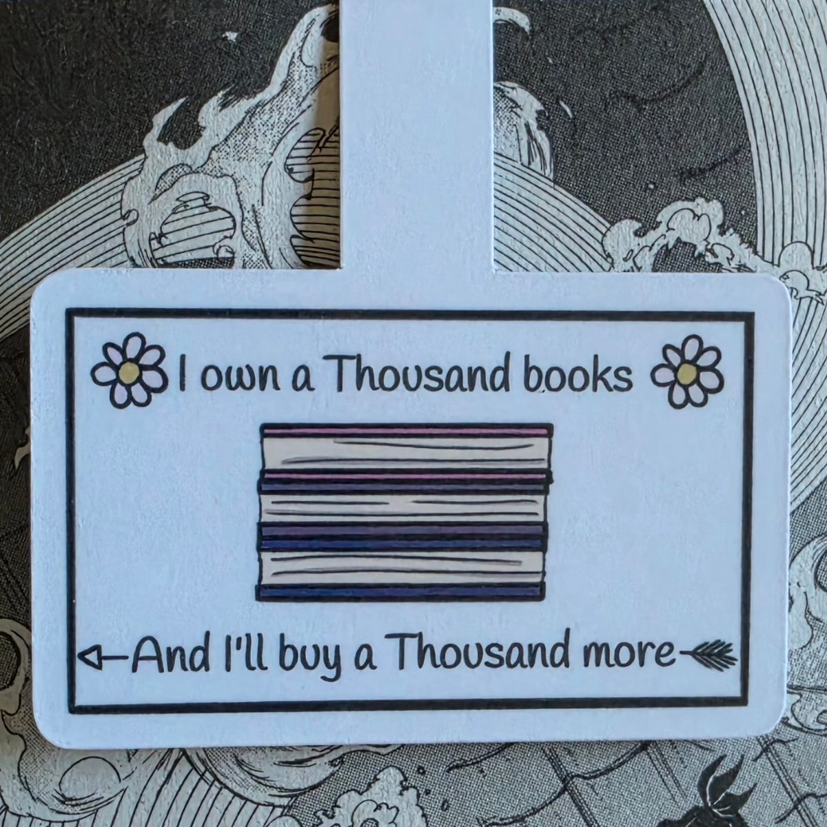 A rectangular magnetic bookmark with rounded corners featuring a printed illustration of stacked books and the text 'I own a Thousand books And I'll buy a Thousand more,' shown against a dark manga illustration background.