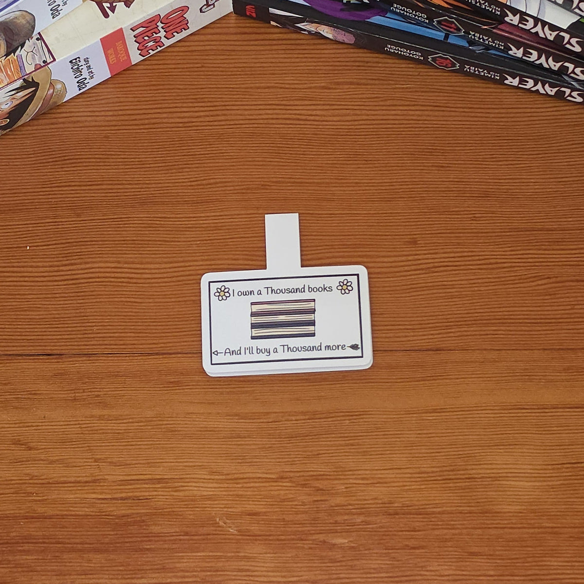 A rectangular magnetic bookmark with rounded corners featuring a printed illustration of stacked books and the text 'I own a Thousand books And I'll buy a Thousand more,' on a wooden table with manga books stacked in the top-left and top-right corners