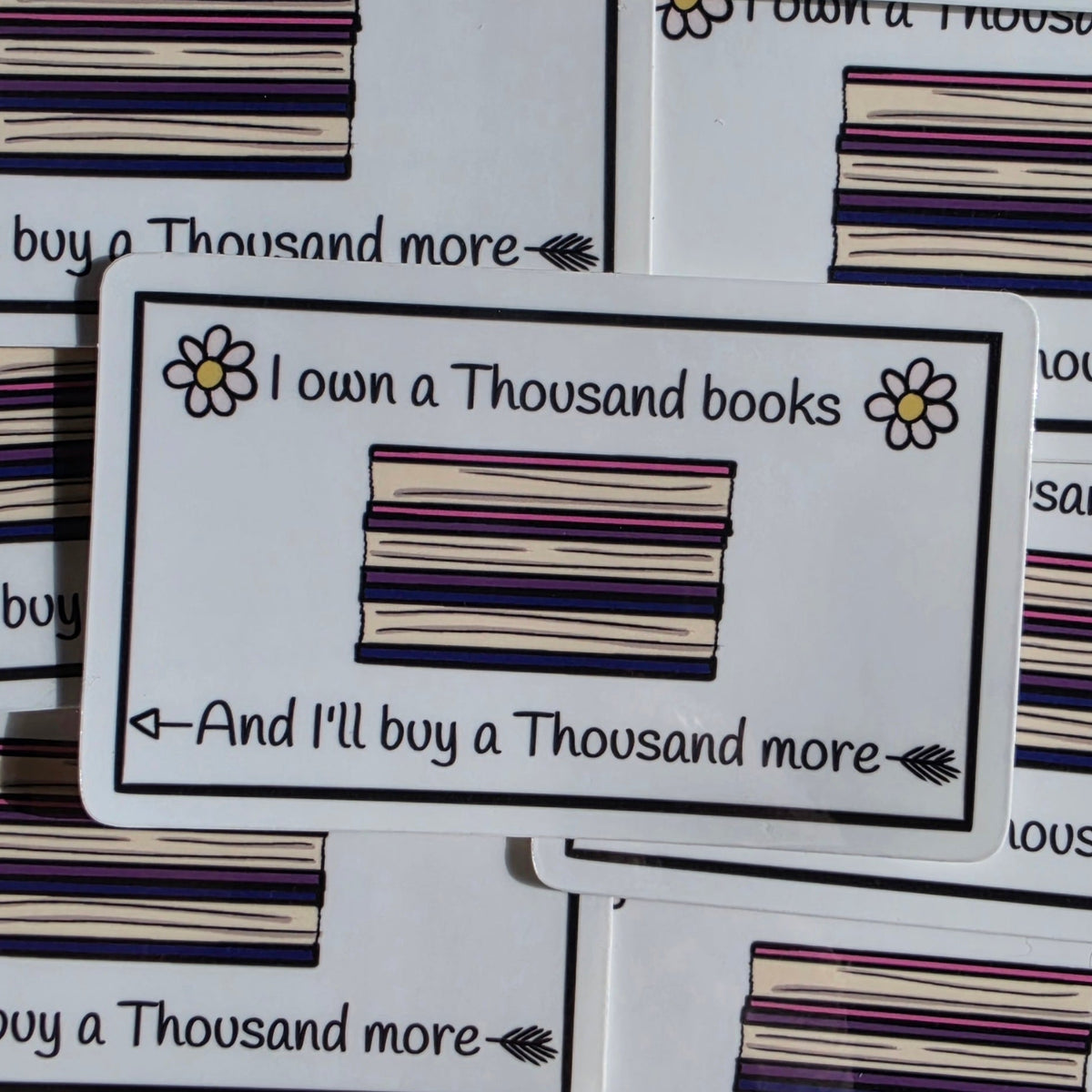 A white rectangular sticker with black text reading 'I own a Thousand books And I'll buy a Thousand more' and a small illustrated stack of books and flowers, surrounded by similar stickers in a pile.