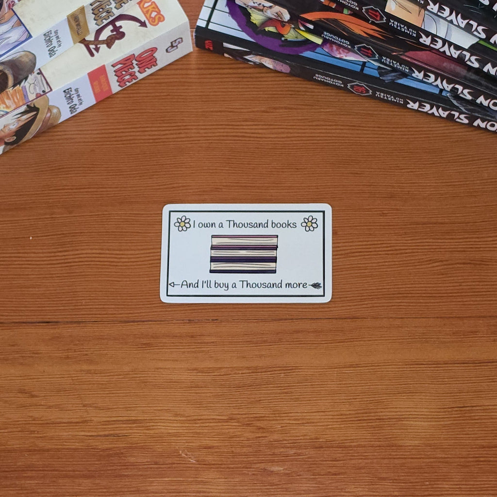 A white rectangular sticker with black text reading 'I own a Thousand books And I'll buy a Thousand more' and a small illustrated stack of books and flowers, on a wooden table with stacks of manga books in the background.