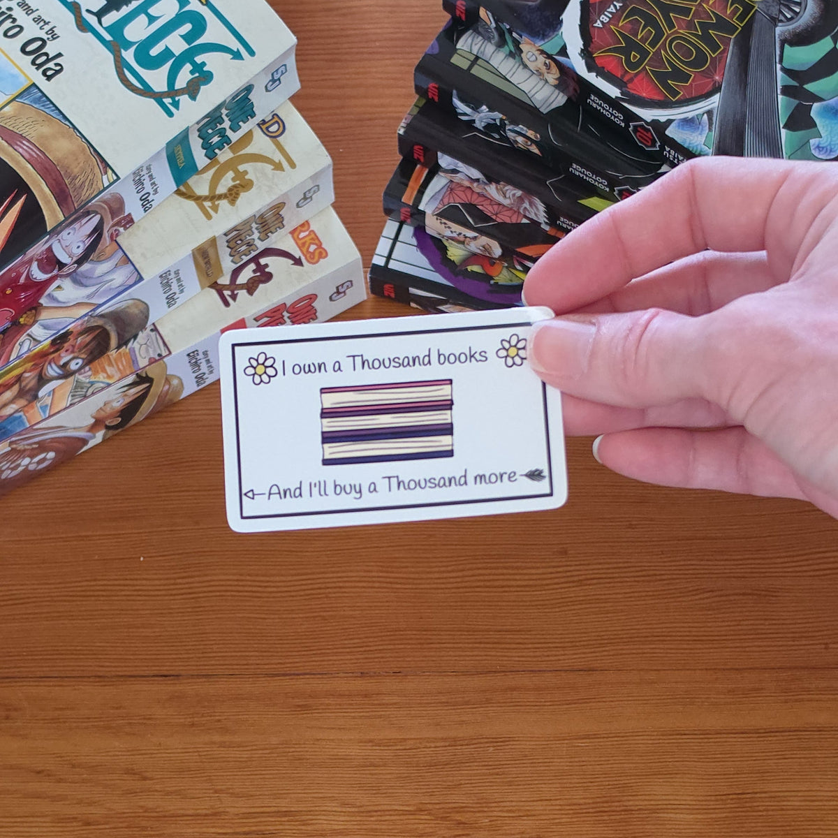 A hand holds a white rectangular sticker with black text reading 'I own a Thousand books And I'll buy a Thousand more' and a small illustrated stack of books and flowers, set against a wooden tabletop with manga books in the background.