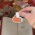 A hand holds a paper craft magnetic bookmark depicting three different colored pumpkins with an open book and autumn decor in the background.