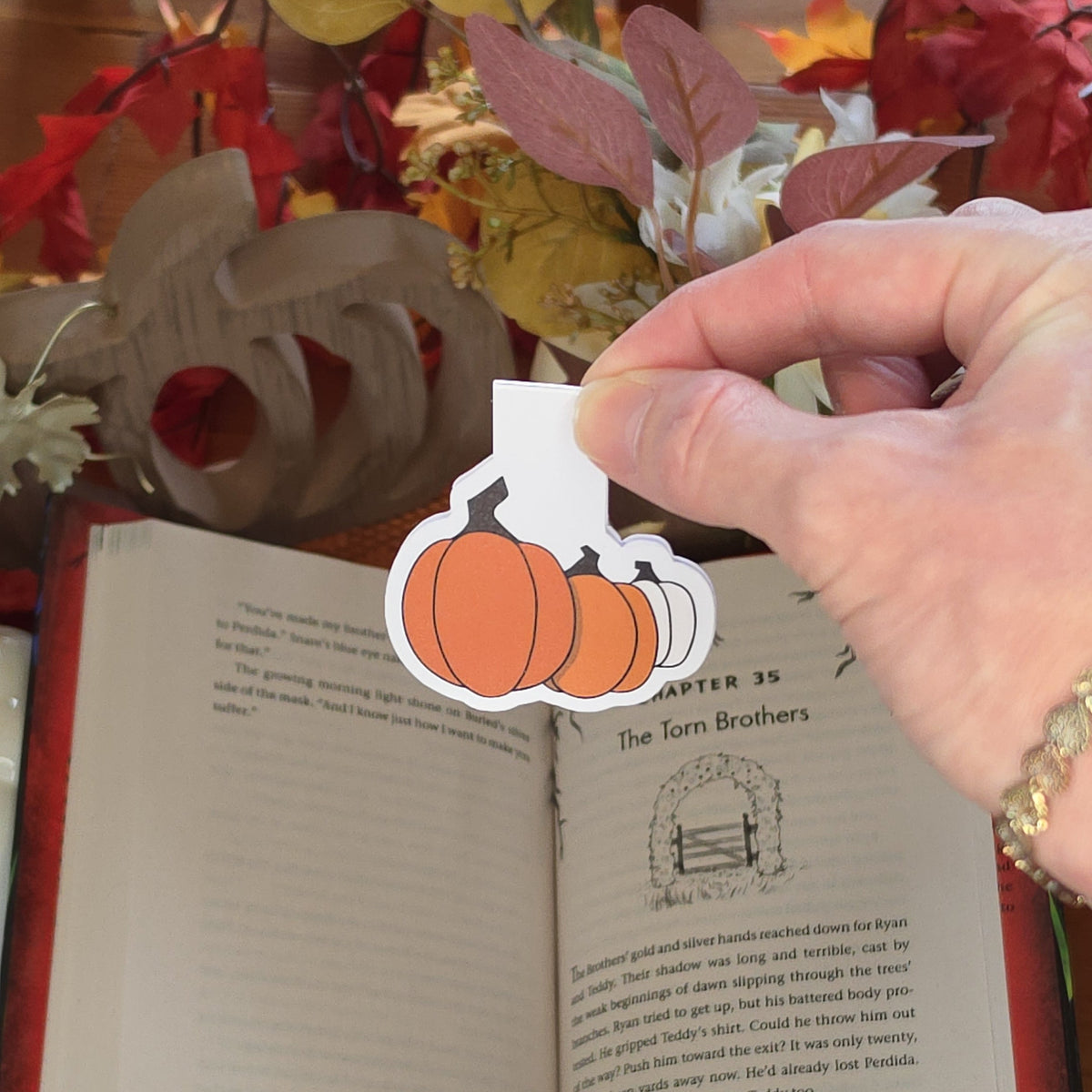A hand holds a paper craft magnetic bookmark depicting three different colored pumpkins with an open book and autumn decor in the background.