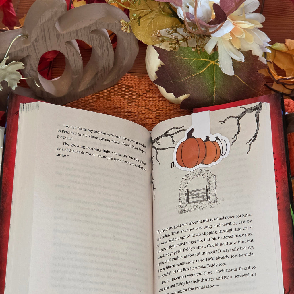 A magnetic bookmark depicting three different colored pumpkins in clasped onto an open book page with autumn decor in the background.
