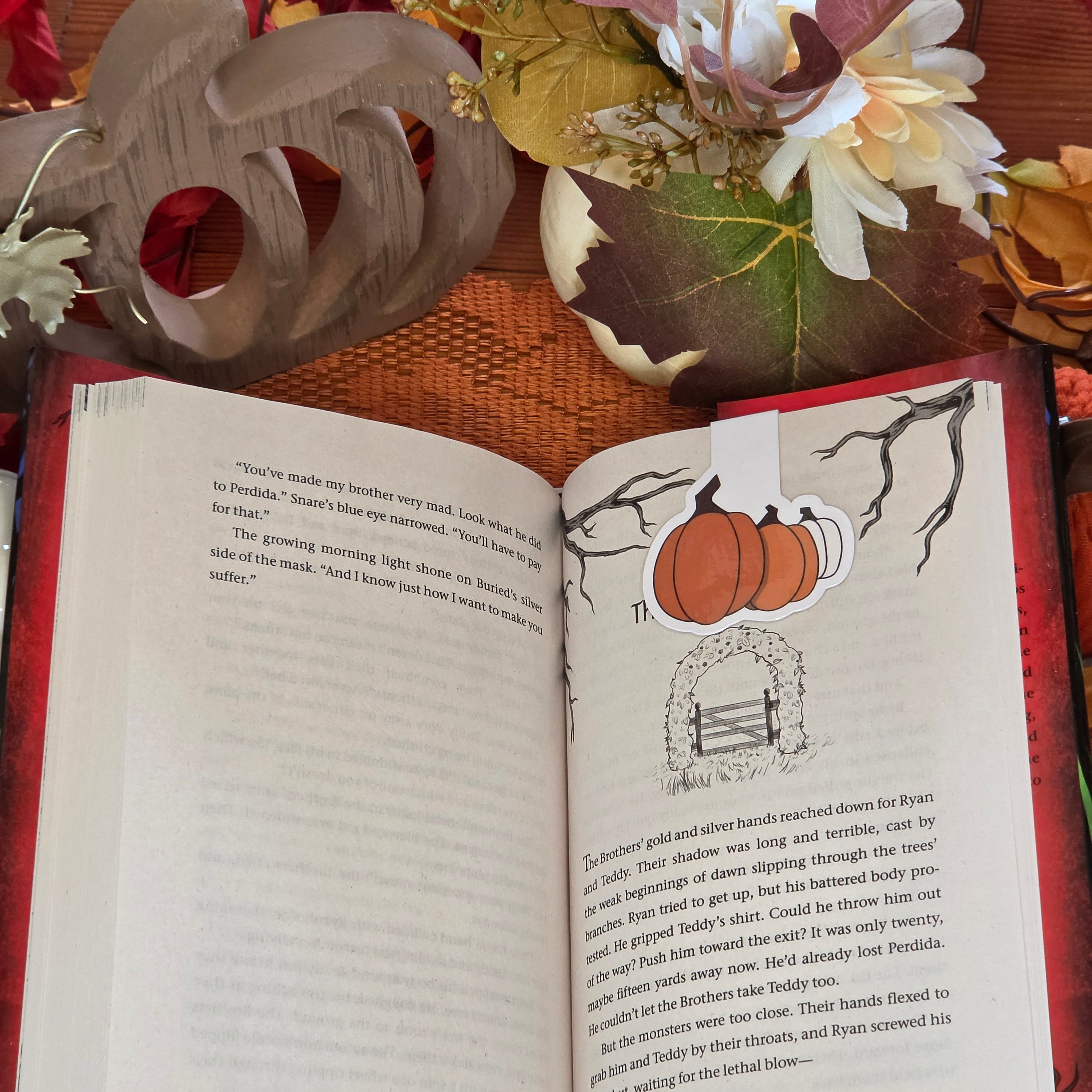A magnetic bookmark depicting three different colored pumpkins in clasped onto an open book page with autumn decor in the background.