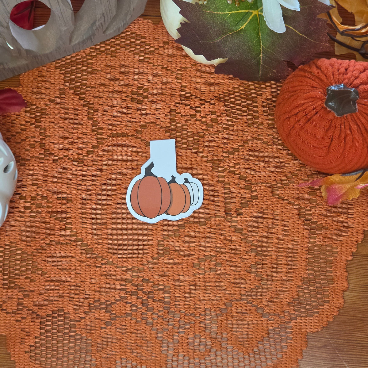 A papercraft magnetic bookmark depicting three different colored pumpkins sits on an orange doily with autumn decor in the background.