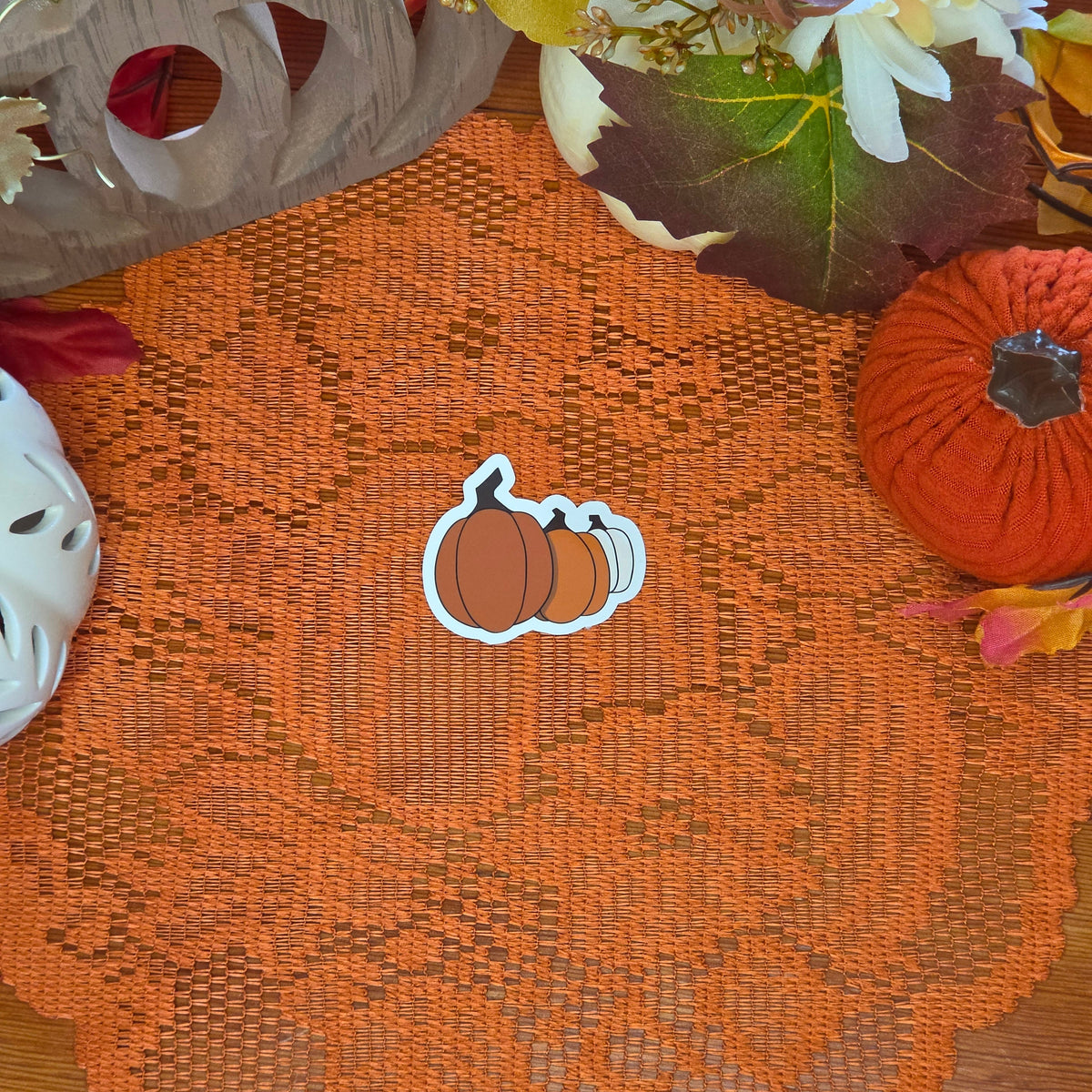 A sticker in the shape of a three pumpkins sticker sitting on an orange doily with autumn decor in the background.