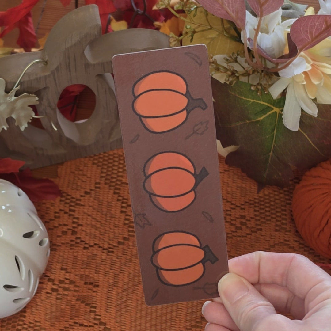 A hand holds a rectangular brown bookmark featuring three orange pumpkin illustrations, set against a cozy autumn-themed background with a lace doily, fall leaves, and pumpkin pottery.
