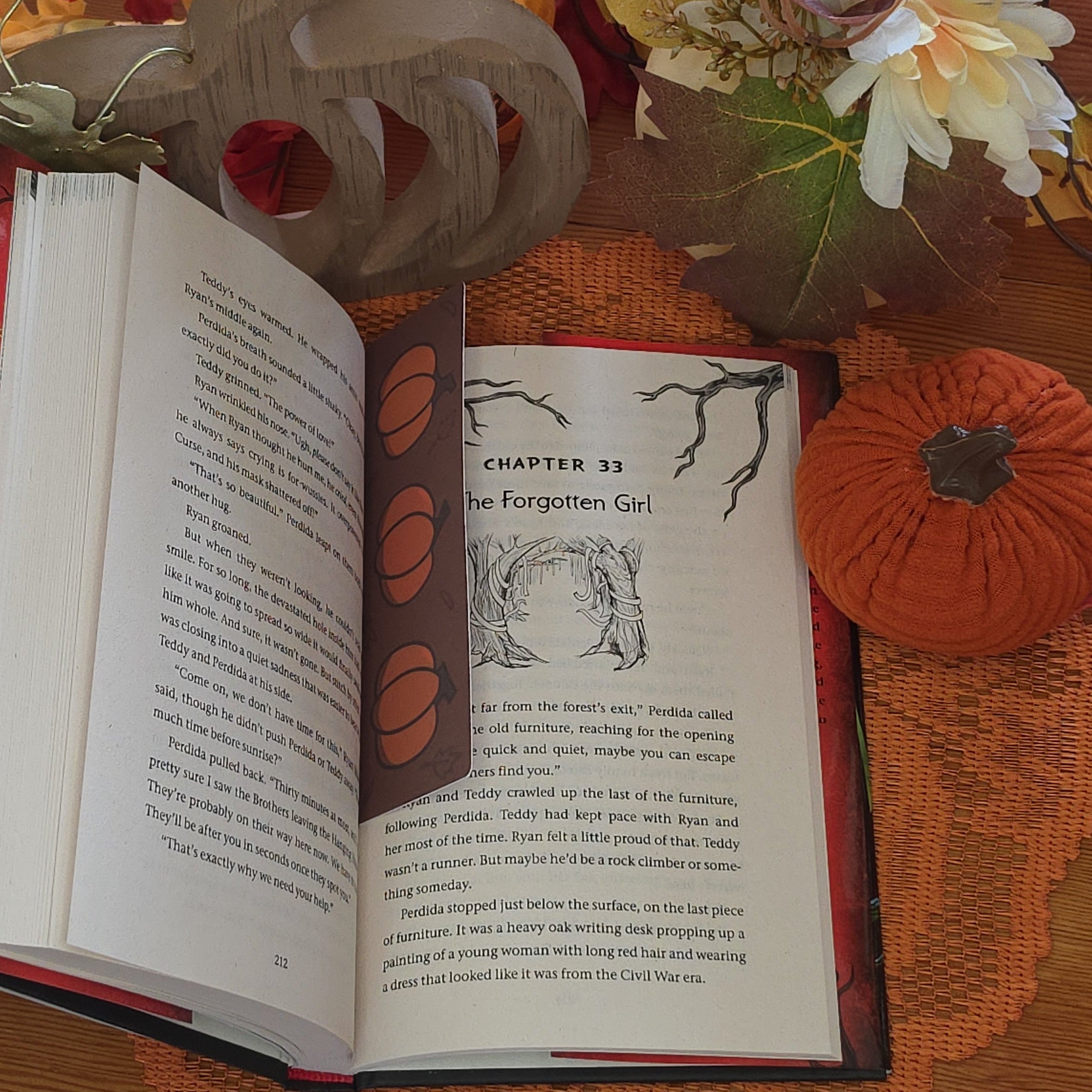 An open book features a rectangular brown bookmark featuring three orange pumpkin illustrations, set against a cozy autumn-themed background with a lace doily, fall leaves, and pumpkin pottery.