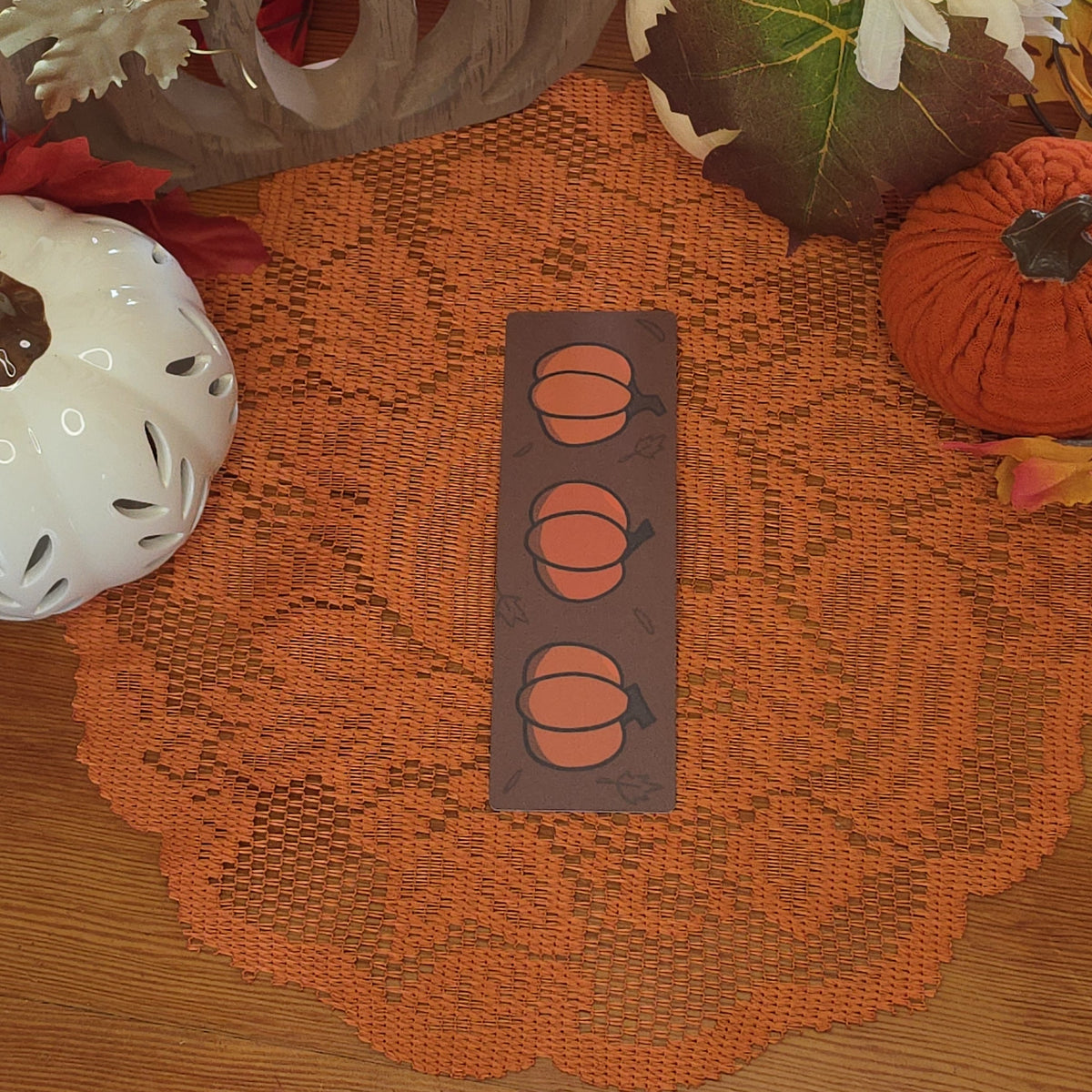 A rectangular brown bookmark featuring three orange pumpkin illustrations, set against a cozy autumn-themed background with a lace doily, fall leaves, and pumpkin pottery.