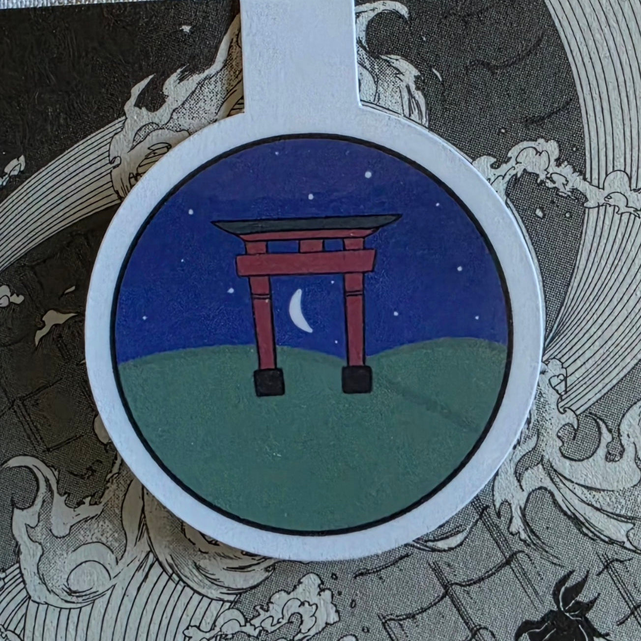 A circular magnetic bookmark featuring a Japanese torii gate at night under a starry blue sky with a crescent moon, shown on a manga illustration.