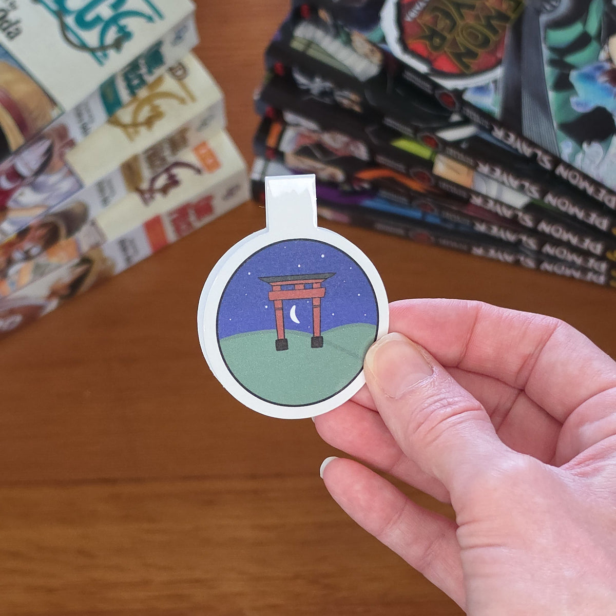 A hand holding a round magnetic bookmark featuring an illustration of a Japanese torii gate at night, with a wooden tabletop and manga books in the background.