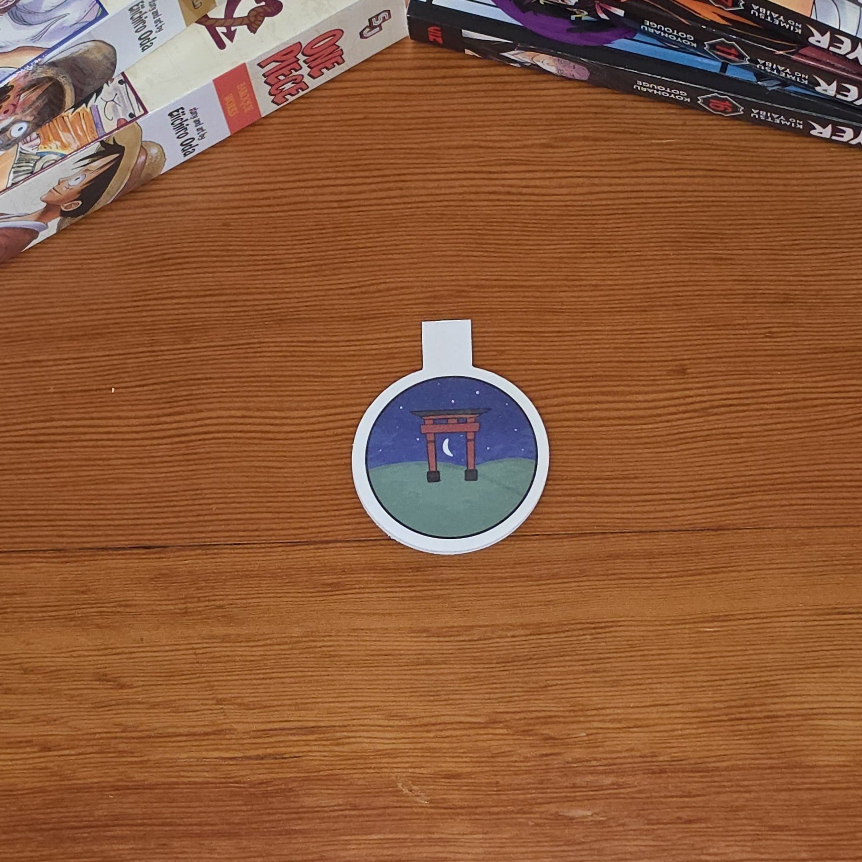 A round magnetic bookmark featuring an illustration of a Japanese torii gate at night, with a wooden tabletop and manga books in the background.