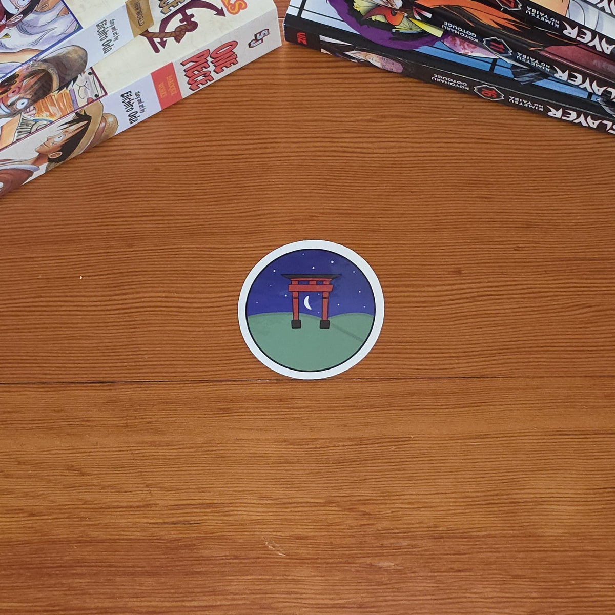 A small round sticker featuring a red torii gate at night under a starry blue sky with a crescent moon, surrounded by a white border illustration on a wooden tabletop, with manga books stacked in the background.
