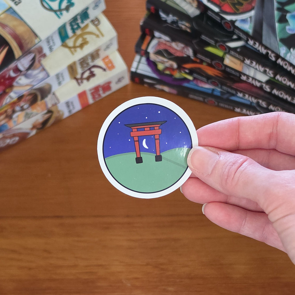 A hand holds a round sticker featuring a red torii gate under a blue night sky on a green hill, with manga books stacked on a wooden table in the background.