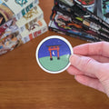 A hand holds a round sticker featuring a red torii gate under a blue night sky on a green hill, with manga books stacked on a wooden table in the background.