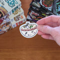 A hand holds a die-cut sticker of a cartoon udon bowl sticker with a winking face with chopsticks, noodles, and toppings in a red-bordered bowl on a wooden table with stacks of manga books in the background.