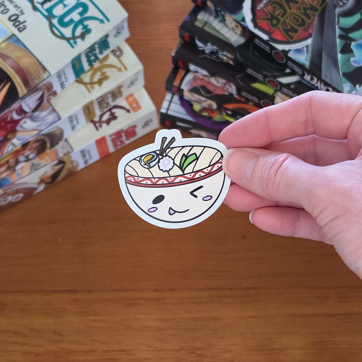 A hand holds a die-cut sticker of a cartoon udon bowl sticker with a winking face with chopsticks, noodles, and toppings in a red-bordered bowl on a wooden table with stacks of manga books in the background.