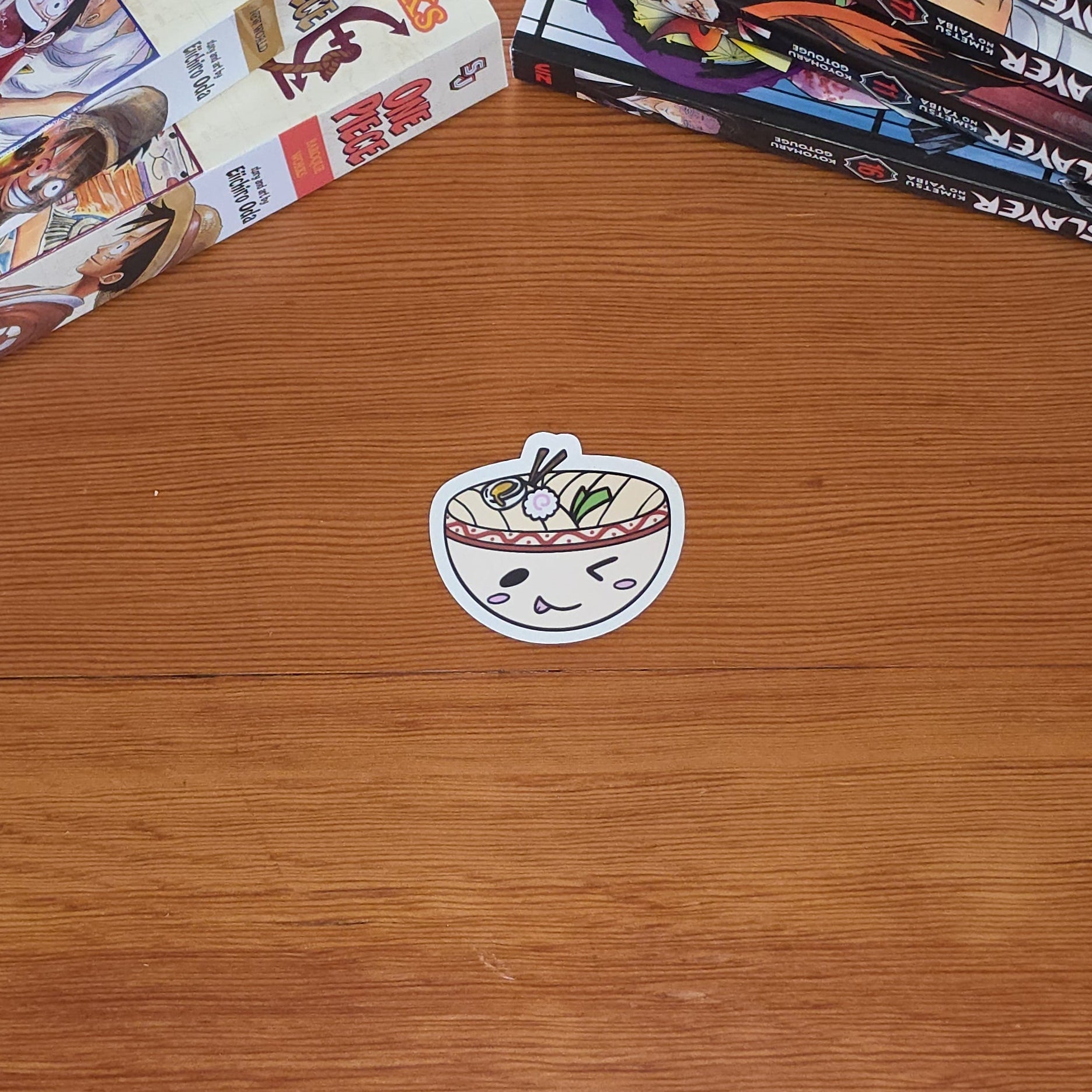 A close-up of a cartoon udon bowl sticker with a winking face with chopsticks, noodles, and toppings in a red-bordered bowl on a wooden table with stacks of manga books in the background.